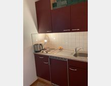 Kleine Küchenzeile mit dunkelroten Schranktüren, einer grauen Arbeitsplatte, einem Edelstahl-Spülbecken und einer zweiflammigen Herdplatte. Die Wand ist mit weißen Fliesen verkleidet, und eine Lampe beleuchtet die Arbeitsfläche.