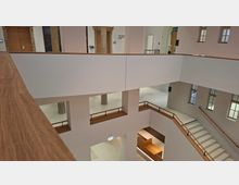 Mehrstöckiges Atrium im Inneren eines öffentlichen Gebäudes mit weißen Wänden, großen Fensteröffnungen und einem Treppenhaus mit Holzhandlauf. Umlaufende Galerien und rechteckige Durchblicke verbinden die Ebenen; unten sind Sitz- und Thekenbereiche aus Holz zu sehen.