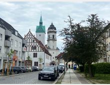 Straßenszene mit gepflastertem Weg, geparkten Autos und einer historischen Kirche mit einem markanten grünen Turm und Giebel im gotischen Stil. Links und rechts der Straße befinden sich mehrstöckige Gebäude, während ein großer Baum den Gehweg auf der rechten Seite säumt.
