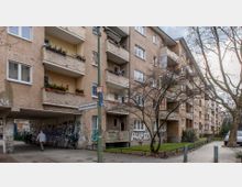 Ein mehrstöckiges Wohngebäude in einem urbanen Umfeld, mit teils verputzter, teils gemauerter Außenfassade und mehreren Balkonen, einige davon mit Pflanzen dekoriert. Im Vordergrund befinden sich ein Gehweg, ein Straßenschild mit der Aufschrift „Hohenstaufenstraße“, sowie ein durch Graffiti gekennzeichneter Durchgang unter dem Gebäude.