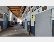 Langer, schmaler Innenraum eines historischen Gebäudes mit weiß gestrichenen Wänden und Holzbalkendecke. An den Wänden sind alte Schilder und Tafeln befestigt, während auf dem Betonboden ein alter Wagen sowie eine rote Waage stehen; die Fenster lassen Tageslicht herein.