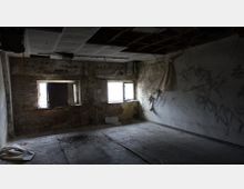 Ein verlassener, heruntergekommener Raum mit abblätternden Wänden, zwei zerbrochenen Fenstern, freiliegenden Ziegeln und teils eingestürzter Decke. Der Boden ist mit Schmutz und Trümmern bedeckt, mit Graffiti an einer der Wände.