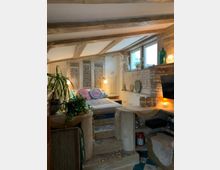 Gemütliches Schlafzimmer mit rustikalem Design, bestehend aus freiliegenden Holzbalken, dekorativen Steinwänden und bunten Fliesen auf den Treppen. Ein erhöhtes Bett mit farbenfrohen Kissen befindet sich neben Fenstern, die Tageslicht hereinlassen, während Pflanzen und warme Beleuchtung eine wohnliche Atmosphäre schaffen.