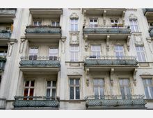 Fassade eines historischen Wohngebäudes mit mehreren Etagen, verziert mit architektonischen Ornamenten und schmiedeeisernen Balkongeländern. Die Fenster und Balkone sind symmetrisch angeordnet, und einige Balkone sind mit Blumenkästen dekoriert.