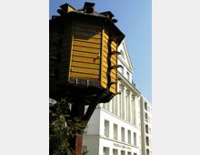 Ein großes, mehrstöckiges Vogelhaus aus Holz mit zahlreichen Öffnungen auf einem hohen Pfosten steht im Vordergrund. Dahinter ist die Fassade der Friedrich-Ebert-Schule mit weißen Wänden und großen Fenstern sichtbar.