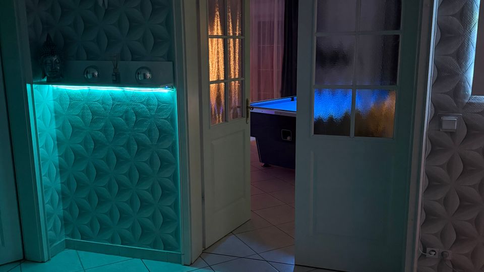 Innenraum mit gemusterten 3D-Wandpaneelen, hellem Fliesenboden und einer halb geöffneten Doppeltür mit Milchglas-Einsätzen. Links steht eine beleuchtete Theke mit türkisfarbenem LED-Licht; durch die Tür ist ein weiterer Raum mit blau beleuchtetem Billardtisch zu sehen.
