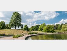Eine Parklandschaft mit einem flachen, geschwungenen Teich im Vordergrund, umgeben von gepflasterten Wegen und Sitzgelegenheiten. Im Hintergrund sind grasbedeckte Hügel mit einzelnen Bäumen und Gruppen von Büschen zu sehen, unter einem teils wolkigen Himmel.