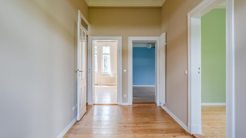 Heller Flur in einer Wohnung mit hohen Decken, Stuckleisten und Holzdielenboden. Mehrere weiße Türrahmen führen in angrenzende Räume, darunter ein blau gestrichener Raum und ein Raum mit grüner Wand sowie ein Fensterbereich am Ende des Flurs.