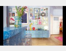 Wohnraum mit hellblauen Wänden und einer bunten Bildergalerie an der Wand. Im Vordergrund eine Theke aus blauem, gesprenkeltem Material mit transparenten Barhockern, im Hintergrund eine Kommode mit Dekorobjekten wie Kerzen und einer Lampe, ergänzt durch helle Holzdielen.