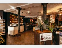 Innenraum einer stilvollen Bar oder eines Restaurants mit langem Tresen, Barhockern und beleuchteten Regalwänden voller Flaschen. Dunkle Säulen, warme Pendelleuchten und Pflanzen rahmen einen Empfangstisch mit „Welcome“-Schild im Vordergrund ein.