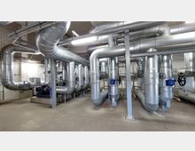 Industrieanlage mit großen, silbernen Rohrleitungen und Ventilen in einem technischen Raum mit Betonboden und weißen Backsteinwänden. Die Leitungen sind teilweise befestigt und verlaufen in komplexen Strukturen durch den Raum.