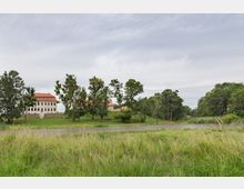 Herrenhaus mit weißer Fassade und rotem Dach inmitten einer weitläufigen, parkähnlichen Landschaft. Im Vordergrund sind eine Wiese und ein kleiner Teich erkennbar, umgeben von Bäumen.