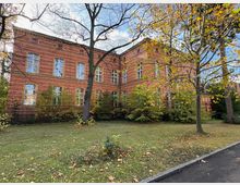 Backsteingebäude im historistischen Stil mit großen Fenstern, umgeben von einer Rasenfläche und Bäumen mit teils herbstlich gefärbtem Laub. Der Himmel ist leicht bewölkt, und ein Gehweg führt entlang des Gebäudes.