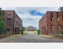 Zweigeschossige Gebäude aus rotem Klinker stehen auf beiden Seiten einer asphaltierten Straße, die auf ein großes, symmetrisches Backsteingebäude zuführt. Im Hintergrund ist das Hauptgebäude mit einer großen Glasfront und dekorativem Steinrelief erkennbar, umgeben von Rasenflächen und niedrigen Hecken.