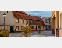Eine historische Gasse mit Kopfsteinpflaster, gesäumt von kleinen, farbigen Häusern mit roten Ziegeldächern und Blumenkästen an den Fenstern. Im Vordergrund stehen Laternen und vereinzelte Bäume mit gelbem Herbstlaub.