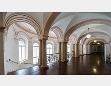 Klassischer Innenraum mit einer Reihe von Rundbögen, die auf dekorativen Säulen stehen, und großen Fenstern mit verziertem Rahmen. Der Boden besteht aus poliertem Holz, und schmiedeeiserne Geländer säumen eine Treppe auf der linken Seite; mehrere Lampen hängen von der gewölbten Decke.