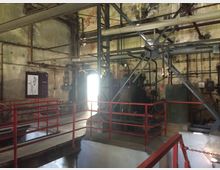 Industriehalle mit verrosteten Metallrohren, Stahlträgern und Maschinen. Rote Geländer umfassen erhöhte Plattformen, und eine gealterte Wand mit technischen Schautafeln ist sichtbar, durchsetzt mit Kabeln und Rohrleitungen.