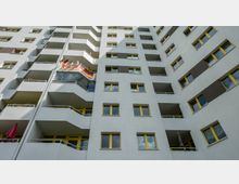 Hohes Wohnhaus mit grauer Fassade, gelb umrahmten Fenstern und Balkonen. Auf einem der Balkone sind mehrere deutsche Flaggen aufgehängt.