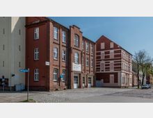 Backstein-Gebäude einer Freiwilligen Feuerwehr mit weißen Toren und Schildern, gelegen an einer gepflasterten Straße. Daneben ein weiteres Gebäude mit roten Ziegeln und verzierten weiß gestrichenen Fassaden.