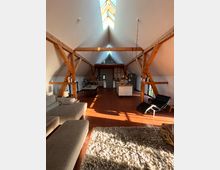 Großzügiger Wohnraum in einem Dachgeschoss mit sichtbaren Holzträgern, großer Dachfensterfront und Terrakotta-Fliesenboden. Im Hintergrund befinden sich eine offene Küche und ein Essbereich, im Vordergrund ein Sofa, ein Teppich und eine Liege.