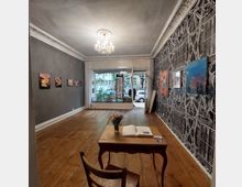 Ein kleiner Kunstgalerie-Raum mit Holzboden und dekorativen Stuckverzierungen an der Decke, beleuchtet von einem Kronleuchter. An den grauen und gemusterten Wänden hängen Gemälde, während ein Tisch mit einem offenen Gästebuch im Vordergrund steht. Im Hintergrund ist eine große Fensterfront mit Blick auf eine Straße zu sehen.