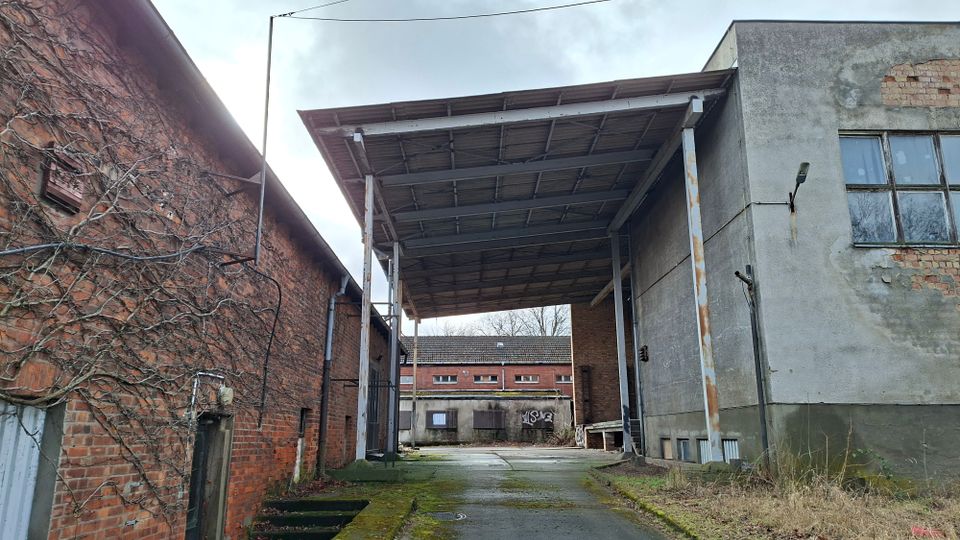 Überdachter Durchfahrtsbereich zwischen einem roten Backsteingebäude und einem grauen Industriegebäude mit großen Fenstern und bröckelndem Putz. Betondecke auf Stahlstützen über einer teils vermoosten Betonfläche; im Hintergrund weitere Betriebsgebäude und eine Mauer mit Graffiti.