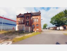 Backsteingebäude mit historischen Fenstern und teilweise zugemauerten Öffnungen in einem Industrie- oder Gewerbegebiet. Daneben befinden sich moderne Lagerhallen und eine asphaltierte Straße mit vereinzelten Bäumen.