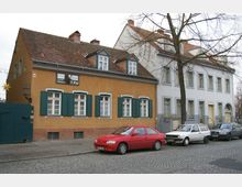 Eine Straßenszene mit einem gelb-braunen historischen Gebäude mit grünen Fensterläden und einem angrenzenden weißen Gebäude mit grauen Fensterrahmen. Mehrere Autos parken entlang der gepflasterten Straße, und ein kahler Baum steht vor den Gebäuden.