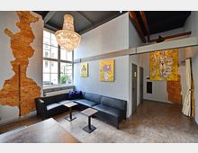 Ein modern gestalteter Innenraum mit einer schwarzen L-förmigen Ledercouch, kleinen Tischen und sichtbaren Backsteinwänden, die teils von hellgrauer Wandfarbe umgeben sind. Große Fenster lassen Tageslicht herein, darüber hängt ein auffälliger Kronleuchter; an den Wänden befinden sich gelbe Gemälde mit abstrakten Motiven.