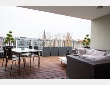 Moderne Terrasse mit Holzboden, ausgestattet mit einem Glastisch und vier Stühlen sowie einem Rattansofa mit Decken. Im Hintergrund eine Balustrade, dahinter eine städtische Wohnanlage und die Spitze eines Kirchturms sichtbar.