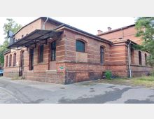 Backsteinbau mit rotem Mauerwerk und gelblichen Zierstreifen, mehreren Rundbogenfenstern und einem Vordach aus Metall. Das Gebäude steht in einer asphaltierten Umgebung mit vereinzelter Vegetation.