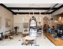 Helles Café mit modernem Interieur, darunter runde Holztische, schlichte Stühle und eine Theke aus Holz auf der rechten Seite. Im Hintergrund sind hohe Bogenfenster, Pflanzen und Regale mit Dekorationen zu sehen, über den Tischen hängen filigrane Pendelleuchten.