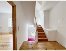 Ein heller Innenraum mit einer Holztreppe, die zu einem oberen Stockwerk führt. Der Raum hat weiß gestrichene Wände, einen Holzboden und Fenster mit weißem Rahmen, die Tageslicht hereinlassen. Links befindet sich ein angrenzender Raum mit Heizkörper und Fenstern.