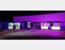 Nachtaufnahme eines Veranstaltungsortes mit beleuchteten Containern als Bars, die in lila und grünem Licht erstrahlen. Im Vordergrund ein sandiger Boden, links und rechts einige Sitzmöglichkeiten, während Menschen vor den Containern stehen.