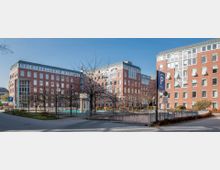 Backsteingebäudekomplex mit mehreren Etagen und großen Fenstern, umgeben von einem gepflasterten Platz mit kahlen Bäumen, Beeten und einer Parkplatzbeschilderung. Im Vordergrund befindet sich eine Tiefgarageneinfahrt mit umlaufendem Geländer.