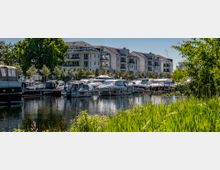 Moderne Wohngebäude mit großen Balkonen im Hintergrund, davor ein Yachthafen mit geparkten Motorbooten auf einem ruhigen Gewässer, umgeben von Bäumen und dichtem Grün im Vordergrund.