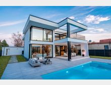Modernes, zweistöckiges Wohnhaus mit Flachdach, großen bodentiefen Glasfronten und überdachter Terrasse mit schwarzen Stützen. Im Vordergrund liegt ein rechteckiger Swimmingpool mit heller Steinfläche, daneben Sitzmöbel; im Hintergrund sind Garten, Zaun und Nachbarhäuser zu sehen.