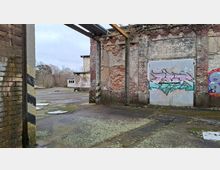 Blick in eine verlassene Industriehalle mit bröckelnden Ziegelwänden, freiliegenden Betonstützen und ohne Dach, sodass der Himmel sichtbar ist. Auf einem Metalltor und der Wand sind großflächige Graffiti, der Boden ist rissig und von Moos sowie Schuttplatten bedeckt, dahinter liegt ein offener Hof mit weiteren Gebäuderesten und Bäumen.