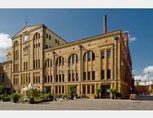 Historisches Backsteingebäude mit gelben Fassaden und großen Rundbogenfenstern, auf dessen Vorderseite "PALAIS" steht. Vor dem Gebäude befinden sich ein gepflasterter Platz, Pflanzen in Kübeln und ein weißes Zelt.