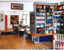 Ein heller Arbeitsraum mit Parkettboden, einer großen Bücherwand voller farbiger Bücher und mehreren Kunstwerken an den weißen Wänden. Im Hintergrund stehen ein Schreibtisch mit einem Computer und ein Bürostuhl vor einem Fenster mit Tageslicht.