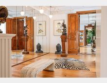 Eingangsbereich eines eleganten Innenraums mit Parkettboden, großen Holz-Doppeltüren und Spiegelwänden. Dekorative Elemente wie eine Buddha-Büste, eine Skulptur auf einem Podest, ein Zebra-Teppich und ein heller Steinhocker verleihen dem Raum eine kunstvolle Atmosphäre.