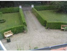 Ein gepflegter Garten mit symmetrischen Rasenflächen, eingerahmt von hohen, geschnittenen Hecken. In der Mitte befindet sich ein Kiesweg, der zu einem schmiedeeisernen Tor führt; links und rechts stehen zwei Holzsitzbänke.