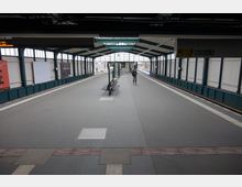 Überdachter Bahnsteig einer U-Bahn-Station mit Glaswänden und Metallstreben, zentral ein Wartebereich mit Sitzbänken und Informationsschildern. Rechts sind Gleise sichtbar, die Beschilderung oben zeigt die Linien U1 und U3 an.