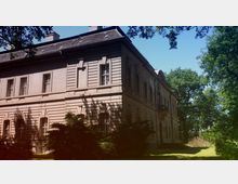 Historisches Gebäude mit einem hellen, detailliert verzierten Fassadenstil und großen Fenstern, umgeben von Bäumen und Schattenwurf auf der Rasenfläche im Vordergrund. Das Gebäude hat ein graues Dach mit Gauben und liegt in einer grünen, parkähnlichen Umgebung.