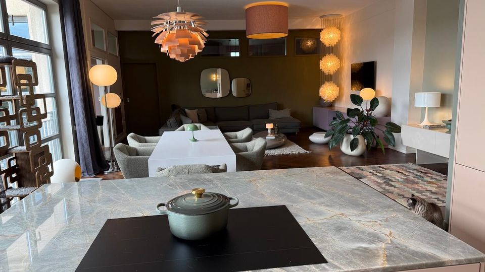 Open-plan modern kitchen and living area with a marble-topped island in the foreground, an induction cooktop, and a dining table leading toward a lounge with a sectional sofa. Large windows, multiple pendant and floor lamps, indoor plants, and a mix of rugs and wood flooring define the space.