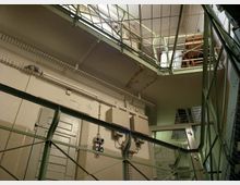 Blick in einen industriellen Innenraum mit Metallgeländern, grünen Stahlträgern und Kabelkanälen. Der Raum enthält eine Tür, elektrische Schaltkästen und ist von mehreren Etagen mit Gitterboden umgeben.