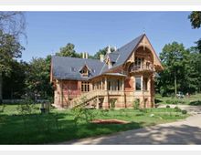 Historische Villa im Park, mit steilem Schieferdach, Ziergiebeln und reich verzierten Holz- und Backsteinfassaden samt Balkon und Veranda. Das Gebäude steht auf einer Rasenfläche mit Wegen und umgebenden Bäumen.