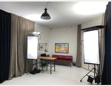 Ein kleines Fotostudio mit weißen Wänden, beige und schwarze Vorhänge, zwei große Softboxen auf Stativen, einem Holztisch in der Mitte und einer roten Ledersitzbank an der Wand. Im Hintergrund hängen gerahmte Bilder.