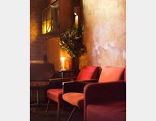 Gemütlicher Innenraum mit einer Wand aus rohem Putz und Ziegeln, roten Polstersesseln, einem Beistelltisch mit Kerze und Blumenarrangement sowie einer Wandlampe. Im Hintergrund ist ein dekoratives altes Metallfenster zu sehen.