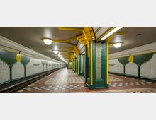 U-Bahn-Station mit rechteckigen Säulen, die mit grünen Fliesen und gelben Metallakzenten verkleidet sind. Die Wände sind mit mosaikartigen Baum- und Blumengrafiken dekoriert, der Boden besteht aus mehrfarbigem Fliesenmuster.